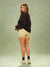 A woman stands in a studio wearing a dark brown sweatshirt, light yellow shorts with small black polka dots, black socks, and white sneakers against a muted green backdrop.