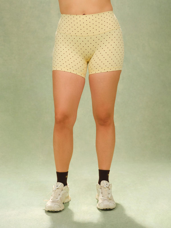 A model's lower body wearing light yellow high-waisted athletic shorts with a small black polka dot pattern, paired with black socks and white sneakers, against a light green studio backdrop.