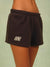 A close-up of a person wearing dark brown sweat shorts with a white embroidered 'SET' logo on the left leg, set against a soft sage green background. <model-info>Meeya wears a size XS and is 5’7, 23” waist, 32B bust.</model-info> 
