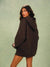 A woman modeling an oversized dark brown hoodie sweatshirt dress against a textured sage green studio background.