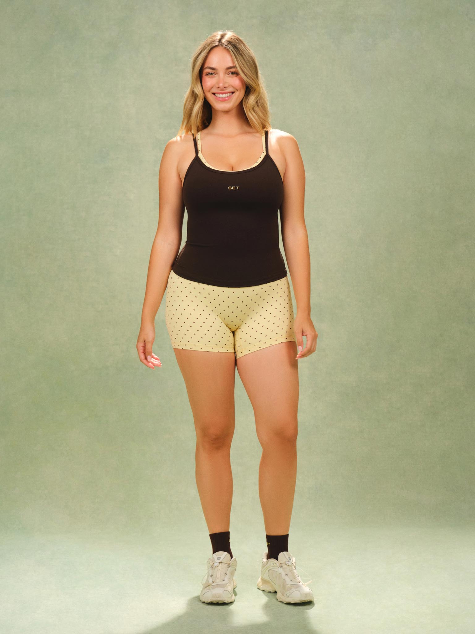A woman modeling a black tank top and light yellow patterned shorts against a textured sage green studio backdrop.