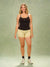 A woman modeling a black tank top and light yellow patterned shorts against a textured sage green studio backdrop.