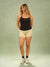 A woman modeling a black tank top and light yellow patterned shorts against a sage green studio backdrop.