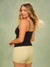 A woman modeling a black camisole-style top with beige trim and matching beige polka-dot shorts against a muted sage green studio background.