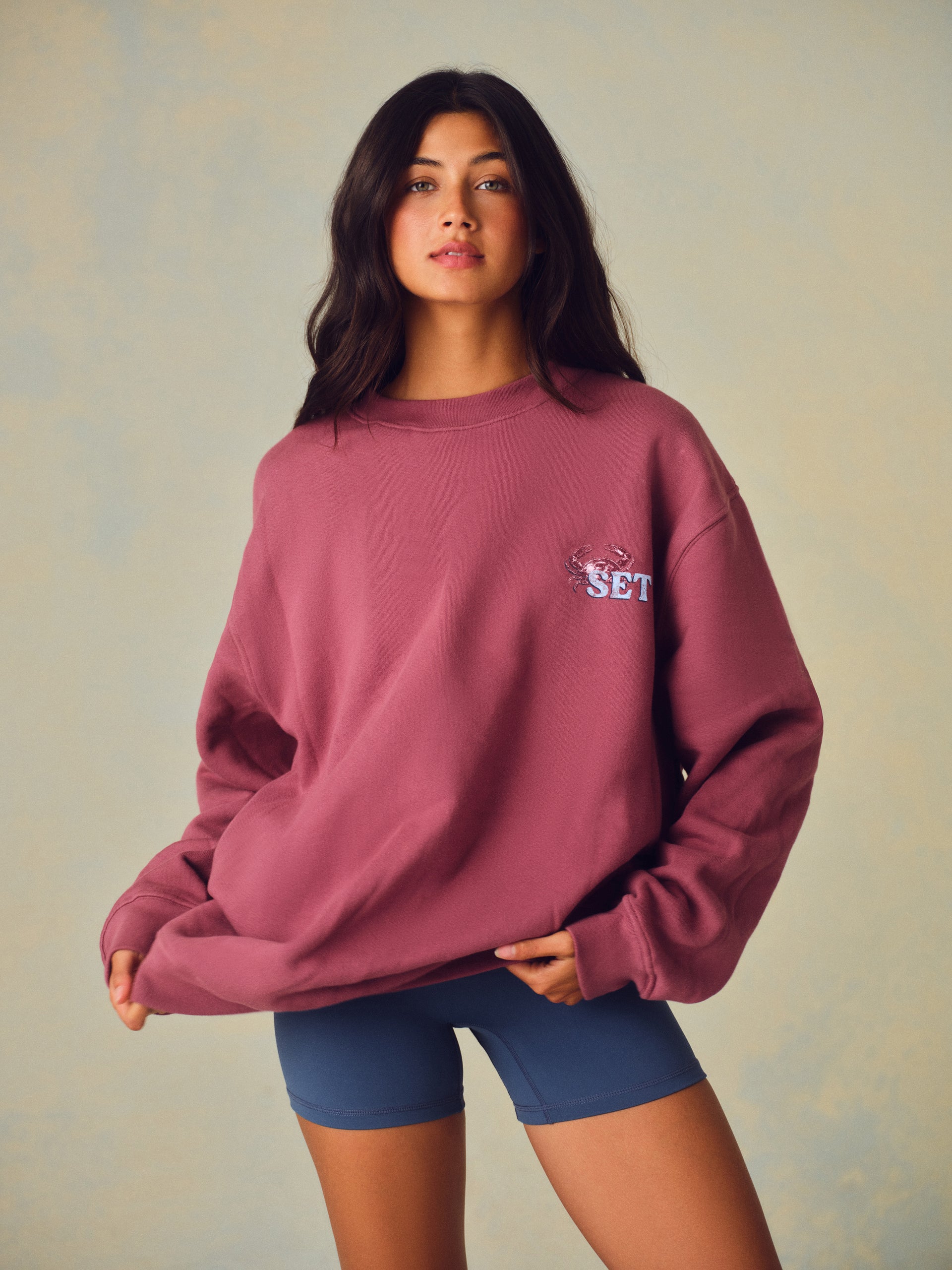 A woman modeling an oversized dusty rose crewneck sweatshirt with a small embroidered 'SET' graphic on the left chest, paired with blue bike shorts against a textured beige studio backdrop. <model-info>Meeya wears a size XS and is 5’7, 23” waist, 32B bust.</model-info> 
