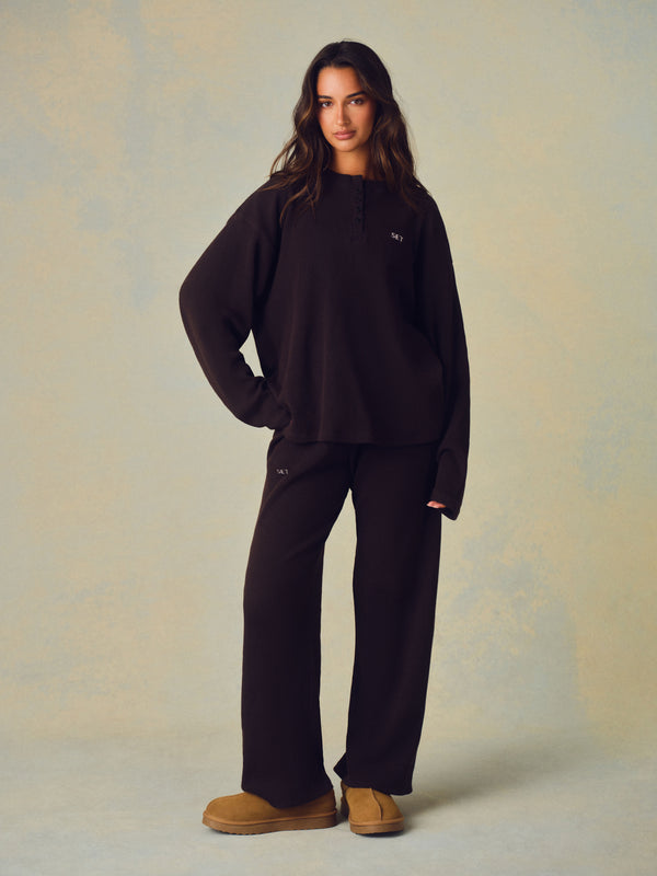 A woman modeling a dark brown lounge set consisting of a long-sleeve mock-neck sweatshirt and matching wide-leg sweatpants, paired with tan suede boots against a textured beige studio backdrop. 