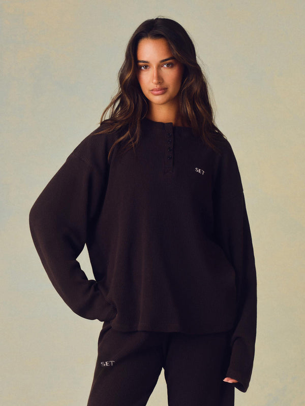 A woman modeling a dark brown long-sleeve henley sweatshirt with a small white embroidered logo on the chest, paired with matching dark pants, against a textured light beige background. 