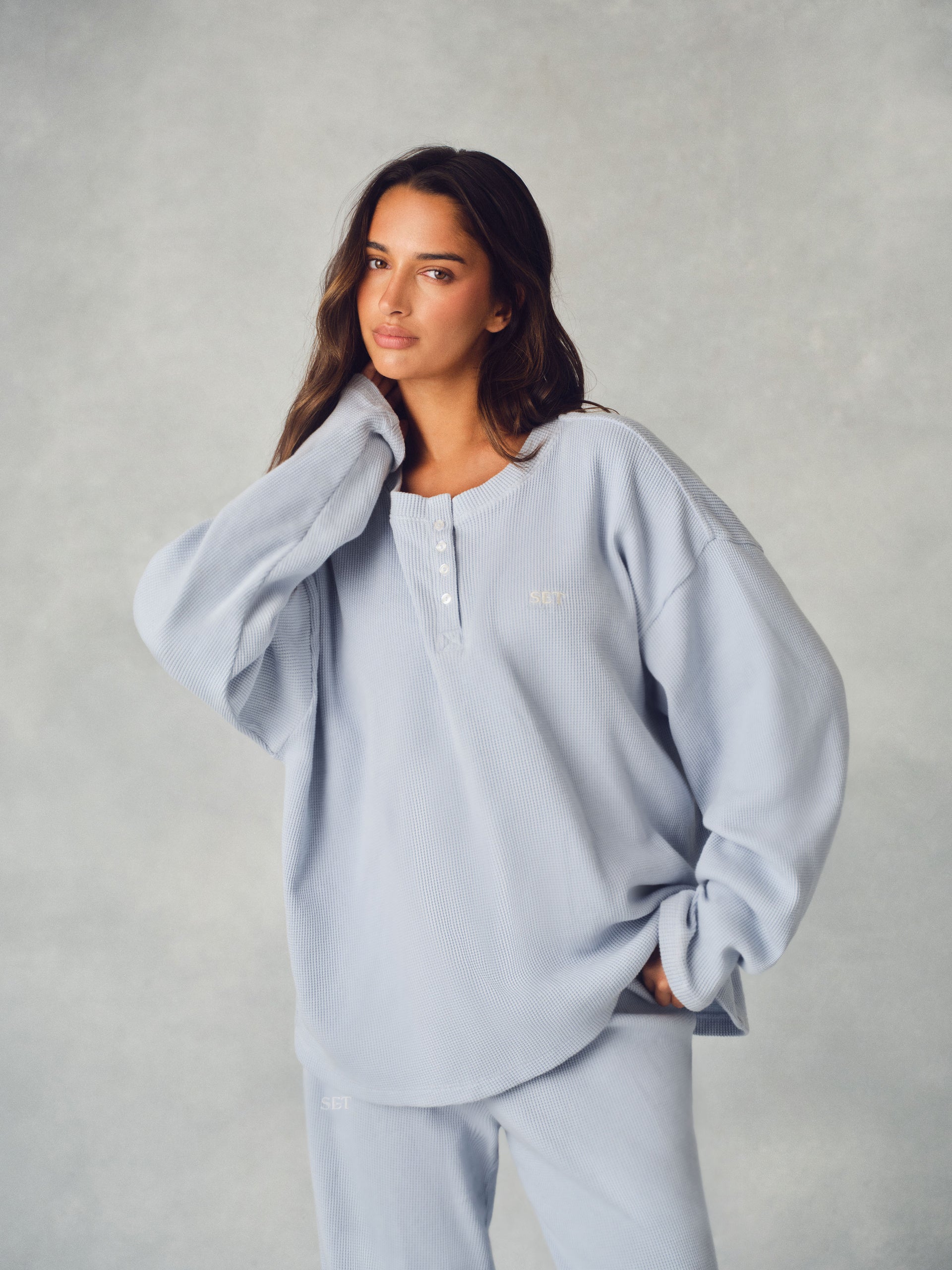 A woman modeling an oversized light blue loungewear set, featuring a long-sleeve henley top with a small white embroidered logo on the chest and matching pants, set against a textured gray studio background. <model-info>Kushi wears a size S and is 5'8, 24” waist, 32C bust.</model-info>