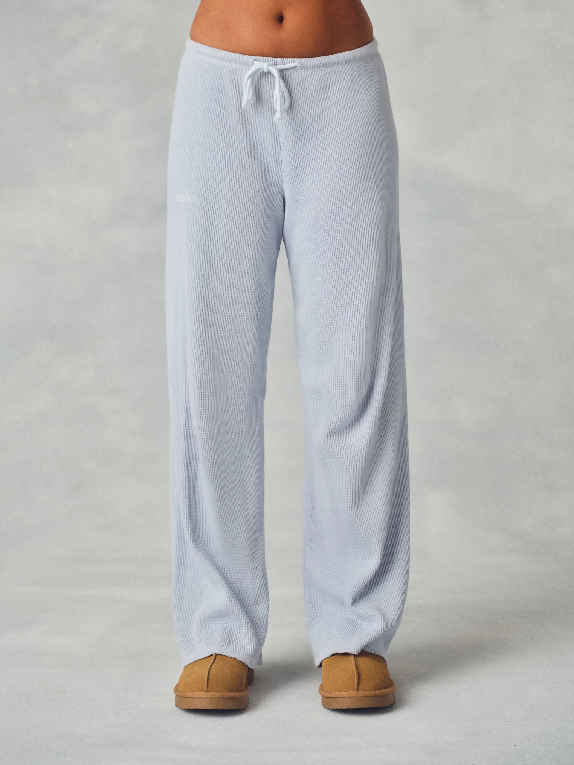 A person wearing light blue wide-leg lounge pants with a white drawstring waist, paired with tan slippers, shown from the waist down against a neutral gray studio background.