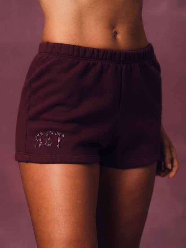 A close-up of a model wearing dark burgundy sweat shorts with a glittery 'SET' monogram on the left leg, set against a solid mauve background. 