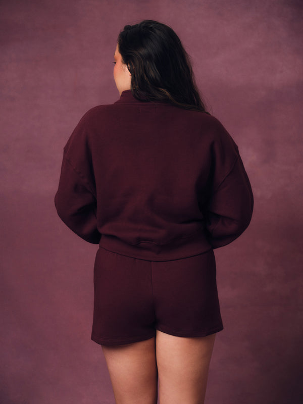 A person shown from the back wearing a matching burgundy sweatshirt and shorts set against a textured mauve studio backdrop.