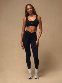 A woman modeling a matching black activewear set consisting of a scoop-neck sports bra and high-waisted leggings, paired with white socks and gray sneakers, against a solid beige studio backdrop. 