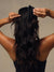A woman viewed from behind with long, wavy black hair, holding a black claw-style hair clip at the top of her head against a plain warm beige background.