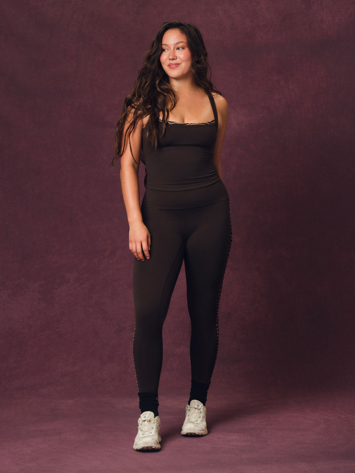A woman modeling a black activewear set consisting of a tank top and matching leggings with side chain detailing, paired with white sneakers, standing against a textured dark purple studio backdrop. <model-info> Christina wears a size L and is 5’9, 29½” waist, 36" bust.</model-info> 