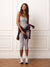 A woman stands indoors against a white paneled wall wearing a light gray ribbed tank top and matching capri leggings, with a dark plum cardigan tied around her waist and white sneakers.
