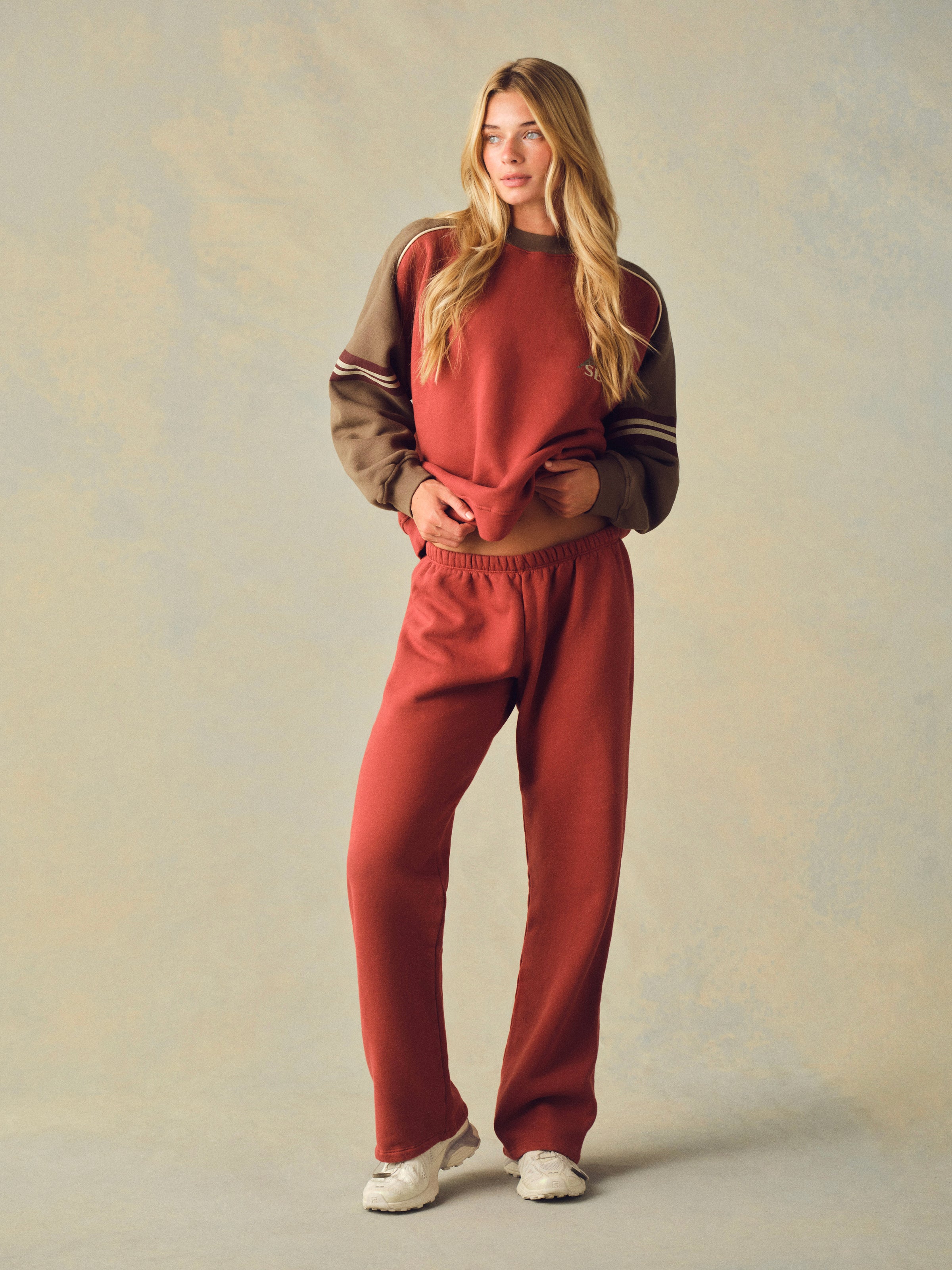 A woman modeling a rust-red sweatshirt and matching sweatpants lounge set against a textured beige studio  