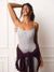 A woman modeling a light gray ribbed bodysuit with thin straps and a small 'BET' logo on the chest, standing against a white paneled wall and wearing a dark purple sweater tied around her waist. <model-info>Meeya wears a size XS and is 5’8”, 23” waist, 32B bust.</model-info>