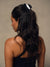 A woman with long, wavy dark hair is shown from the back, wearing a glossy black claw clip holding her hair at the top. She is wearing a black spaghetti strap top against a neutral, textured beige background.