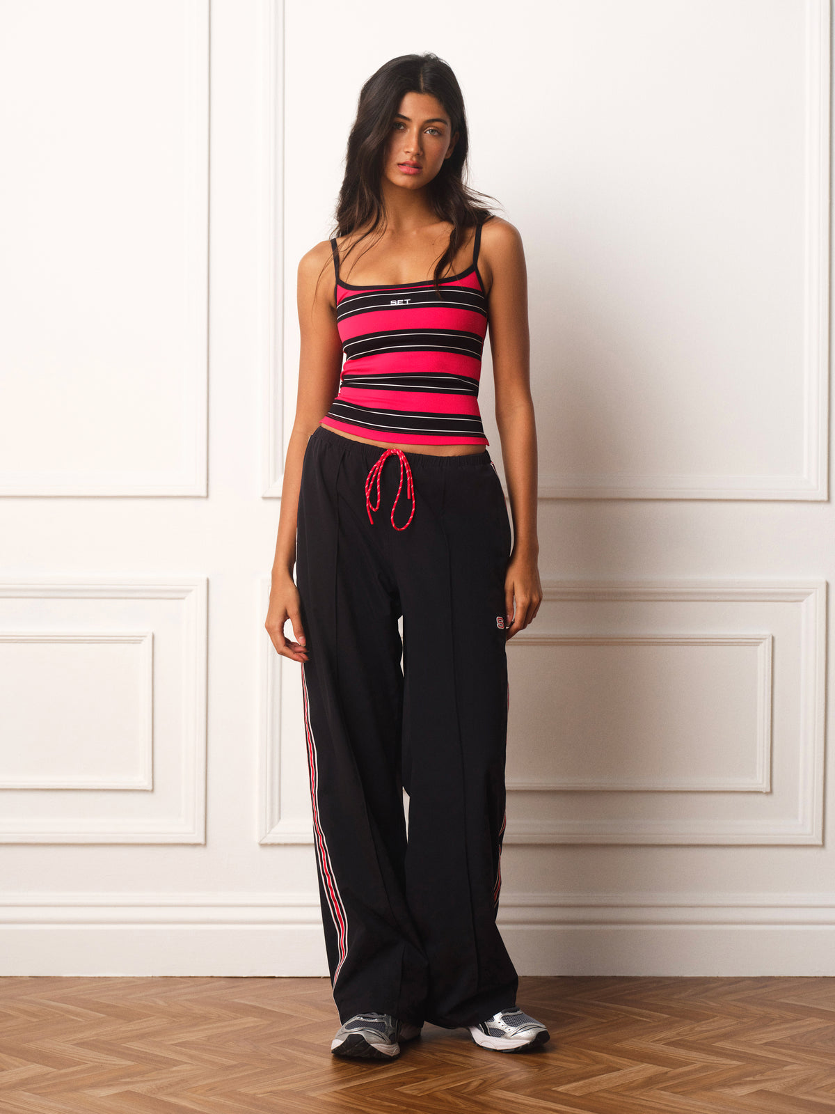 A woman stands against a white paneled wall wearing a black camisole top with bright red and black horizontal stripes, paired with black wide-leg sweatpants featuring red side stripes and a red drawstring, with gray sneakers.