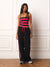 A woman stands against a white paneled wall wearing a black camisole top with bright red and black horizontal stripes, paired with black wide-leg sweatpants featuring red side stripes and a red drawstring, with gray sneakers.
