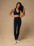A woman modeling a black sports bra and matching high-waisted black leggings against a solid beige studio backdrop, wearing white sneakers.