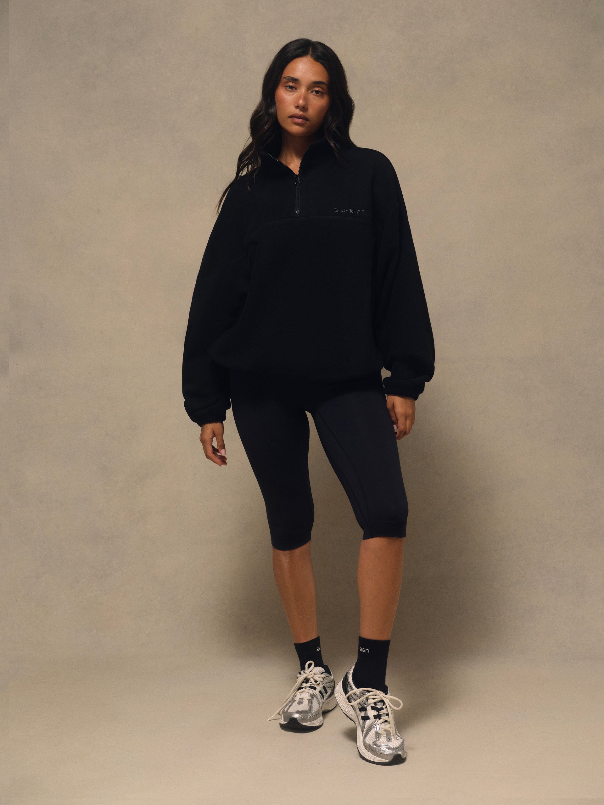 A model stands against a textured beige studio backdrop wearing a black half-zip pullover, black bike shorts, black socks, and white and black sneakers.