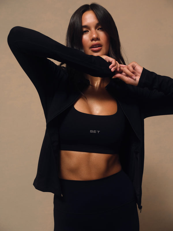 A woman modeling a black athletic set consisting of a sports bra with 'SET' printed on the chest, a matching zip-up jacket, and high-waisted leggings, posed against a solid beige background.