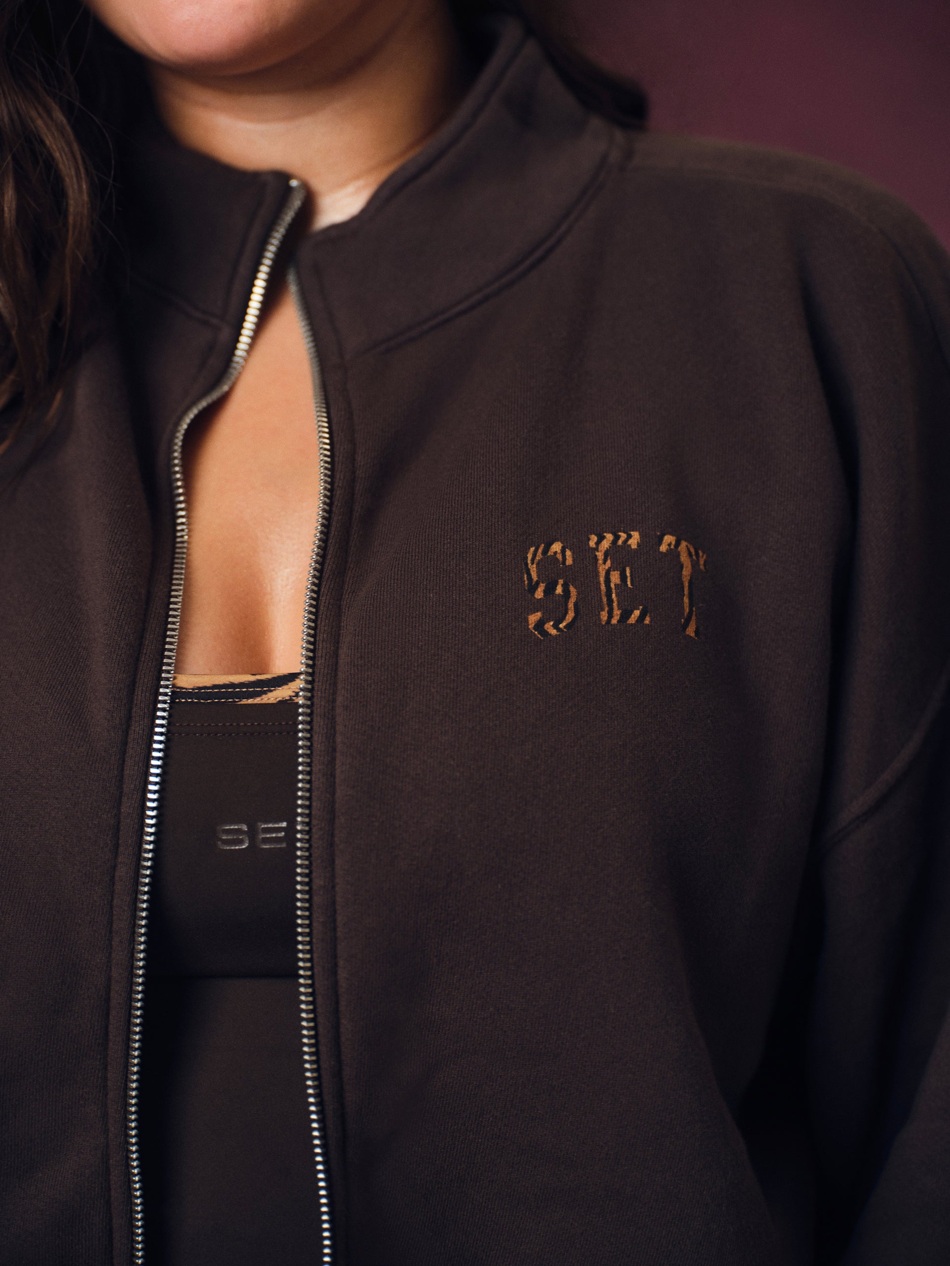 Close-up of a person wearing a dark brown zip-up jacket with a small tiger pattern logo on the chest, layered over a black top.