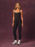 A woman modeling a black athletic tank top and matching black leggings with gold chain-like side detailing, standing against a textured mauve-purple studio backdrop.<model-info> Andrea wears a size S and is 5’9”, 25½” waist, 32” bust.</model-info>