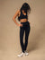 A woman modeling a black athletic sports bra and matching high-waisted leggings, paired with white sneakers, standing against a solid beige studio backdrop. <model-info>Jelene wears a size XS and is 5’7”, 23” waist, 32AB bust.</model-info>