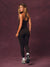 A woman modeling a black athletic jumpsuit with gold chain trim and side details, shown from the back against a textured dark purple studio backdrop.