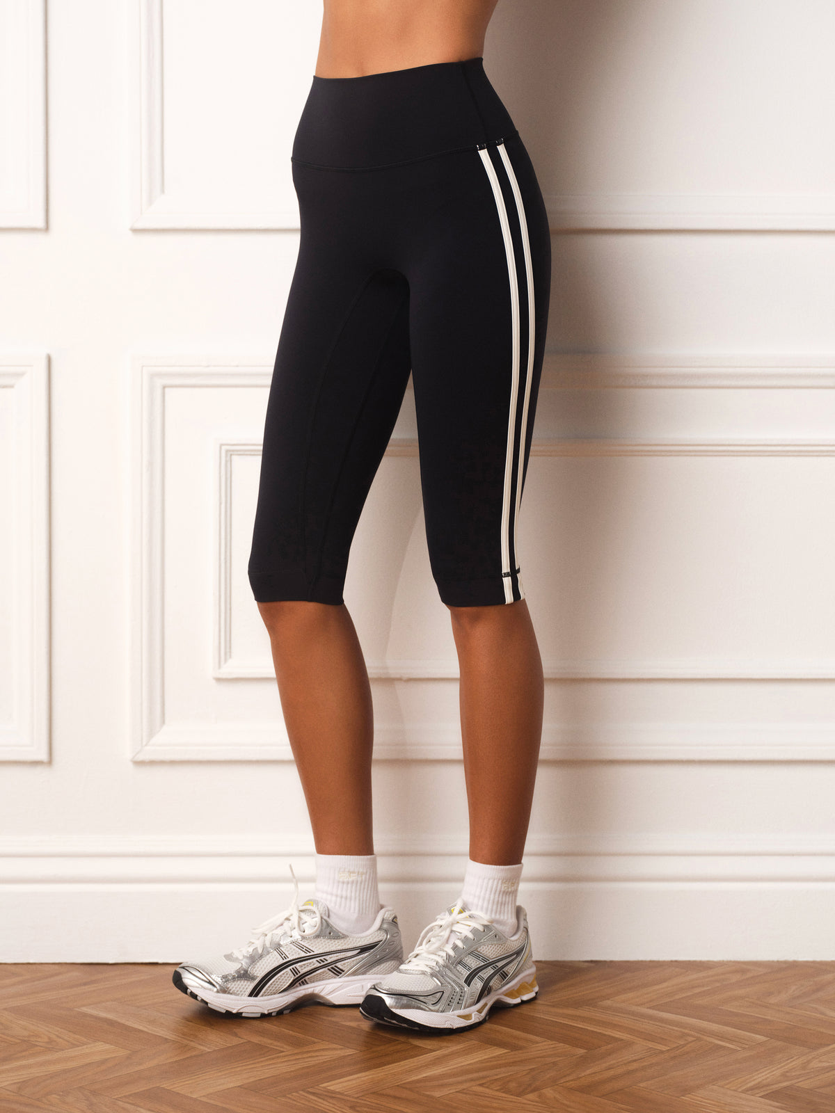 Black ribbed biker shorts with double white side stripes, modeled on a person standing in front of a white paneled wall on a wood floor.