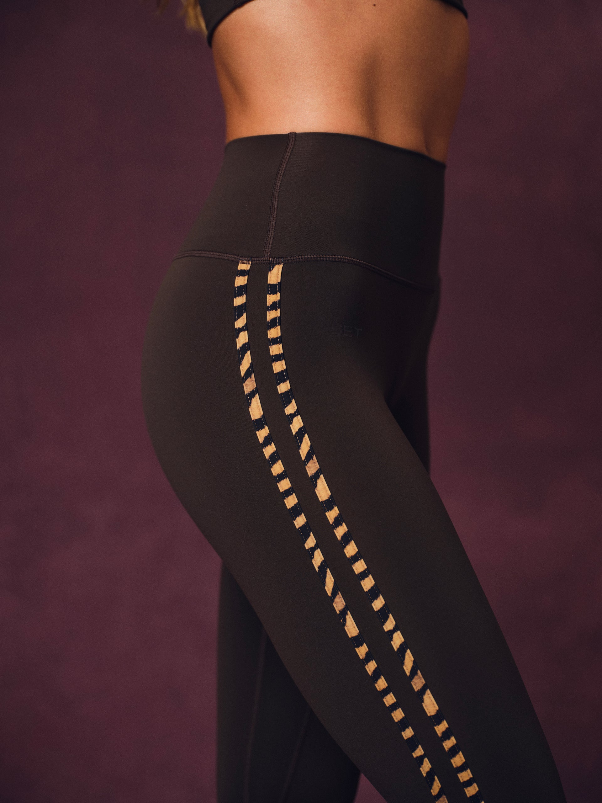 Close-up of a model wearing black high-waisted leggings with gold geometric stripe details down the sides, set against a dark purple background.