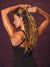 A woman with long highlighted brown hair is shown from the back, wearing a black sports bra and pulling a chunky gold claw clip into her hair against a dark purple studio background.