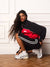A woman crouches in a white paneled room wearing a black and red track jacket, black track pants with white stripes, white socks, and white sneakers.