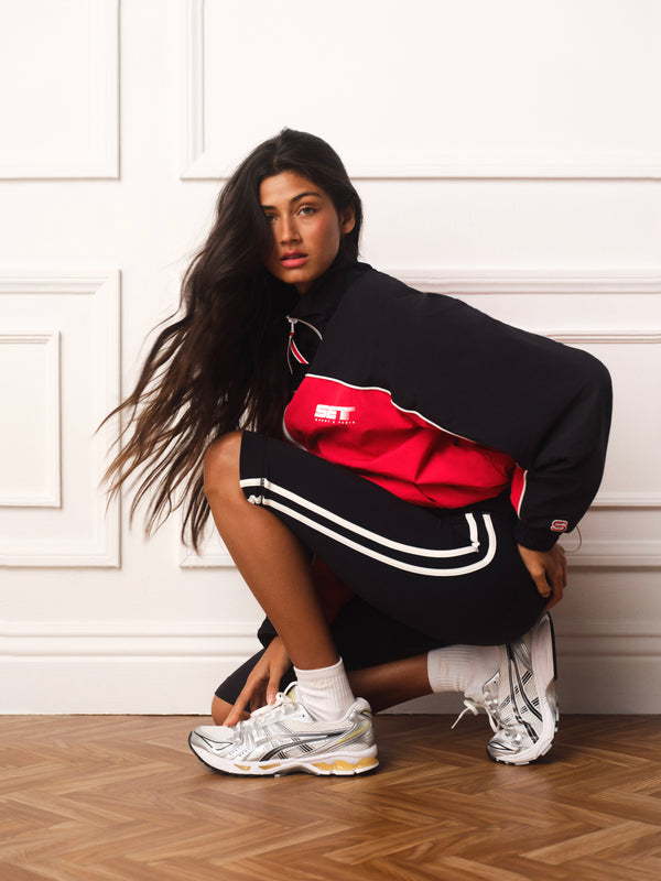 A woman crouches in a white paneled room wearing a black and red track jacket, black track pants with white stripes, white socks, and white sneakers.