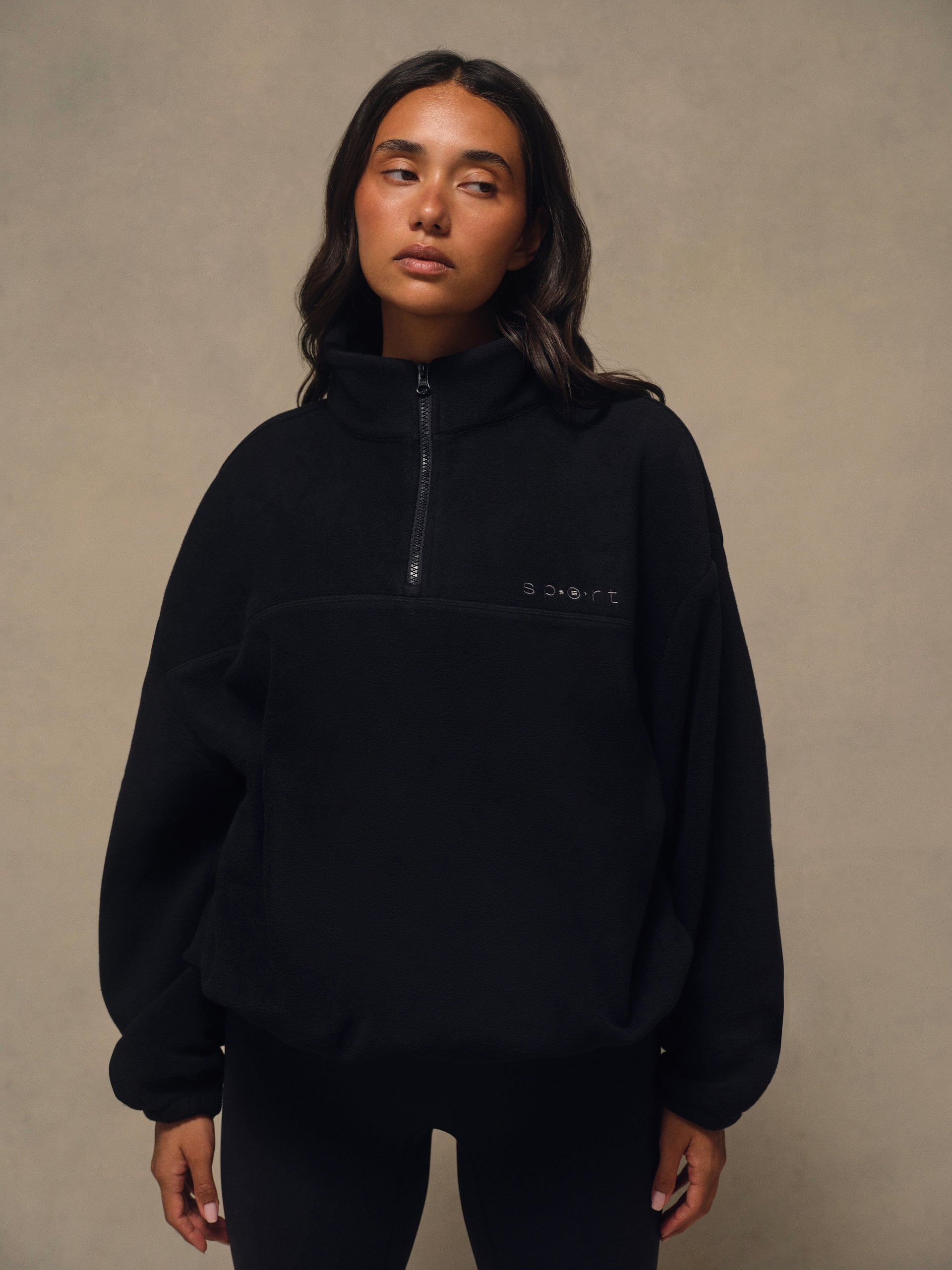 A woman wearing a black fleece pullover with a half-zip neckline and a small white logo on the left chest, standing against a textured beige studio backdrop. <model-info>Irene wears a size S and is 5’8”, 30” waist, 36" bust.</model-info>