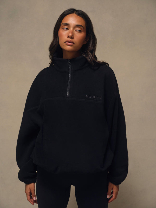 A woman wearing a black fleece pullover with a half-zip neckline and a small white logo on the left chest, standing against a textured beige studio backdrop. 