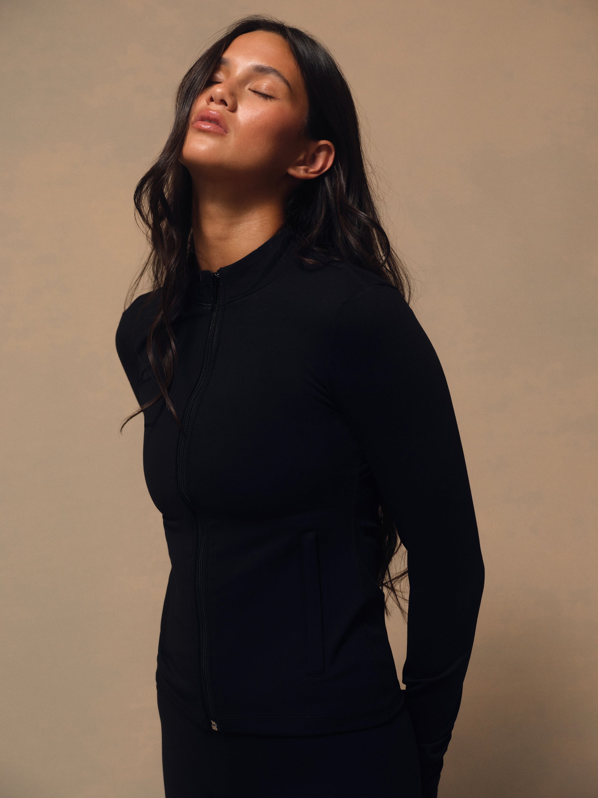 A woman wearing a black long-sleeve athletic zip-up jacket, shown in a side profile against a warm beige studio background. <model-info>Jelene wears a size XS and is 5’7”, 23” waist, 32AB bust.</model-info>