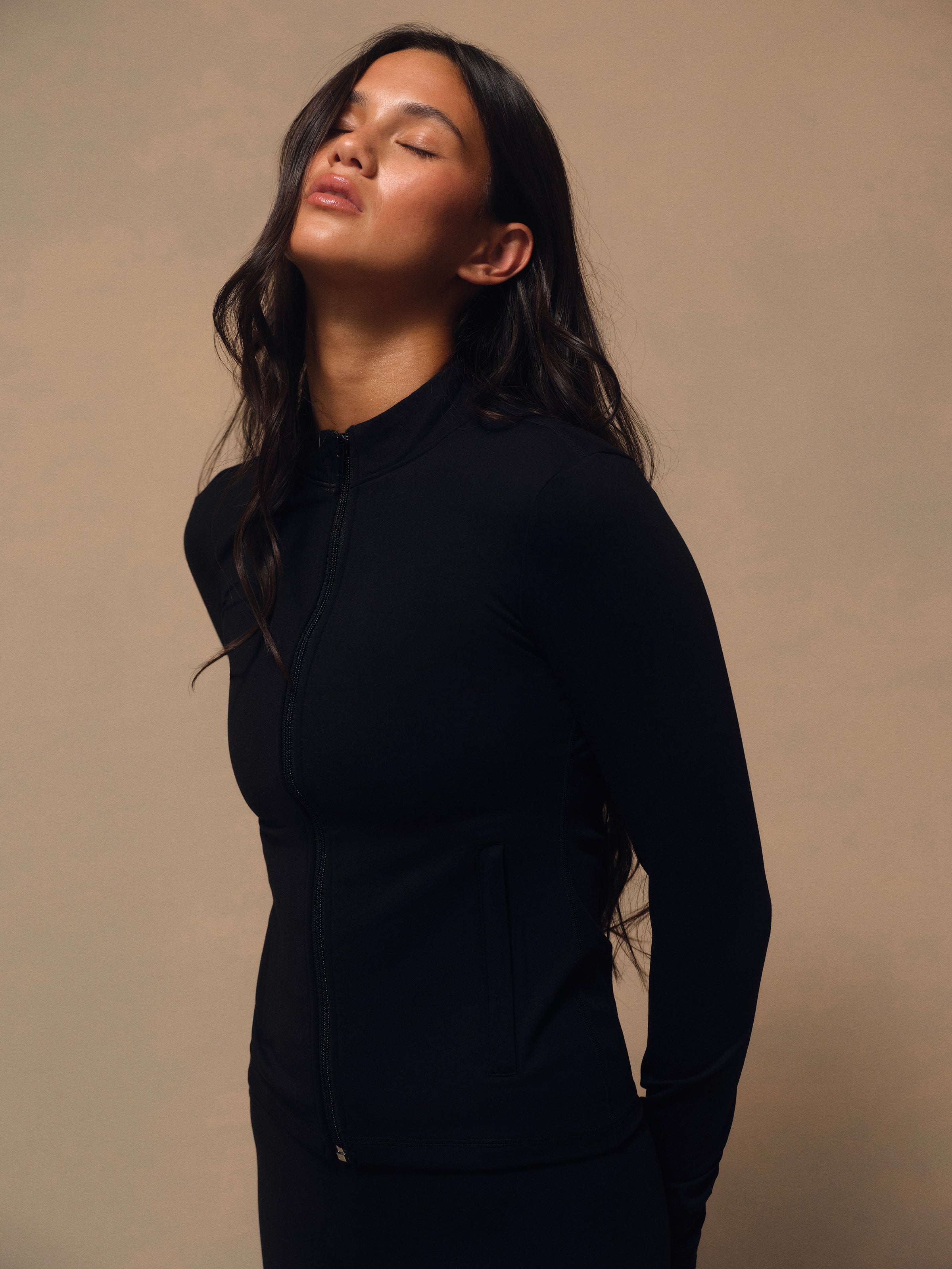 A woman wearing a black long-sleeve athletic zip-up jacket, shown in a side profile against a warm beige studio background. 