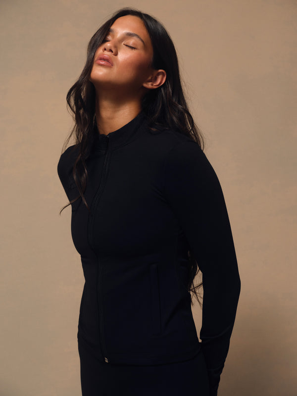 A woman wearing a black long-sleeve athletic zip-up jacket, shown in a side profile against a warm beige studio background. 
