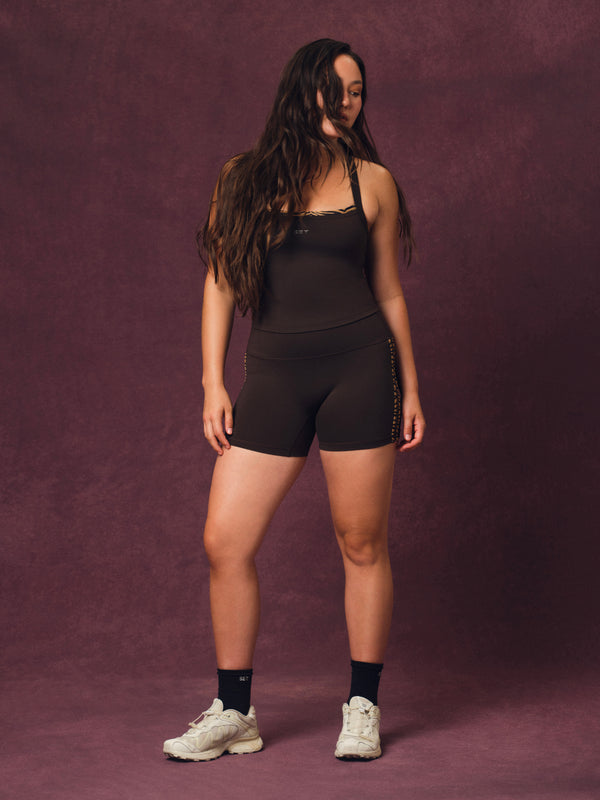 A woman modeling a black athletic set and matching high-waisted shorts, paired with black socks and white sneakers, set against a textured dark purple studio backdrop.