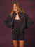 A woman modeling a dark brown zip-up bomber jacket with gold 'SET' embroidery on the chest, paired with matching black activewear, against a textured dark purple studio backdrop.<model-info> Andrea wears a size S and is 5’9”, 25½” waist, 32” bust.</model-info>