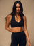 A woman modeling a black sports bra and matching black leggings against a warm beige studio background. The sports bra has a small white 'SET' logo on the underband.