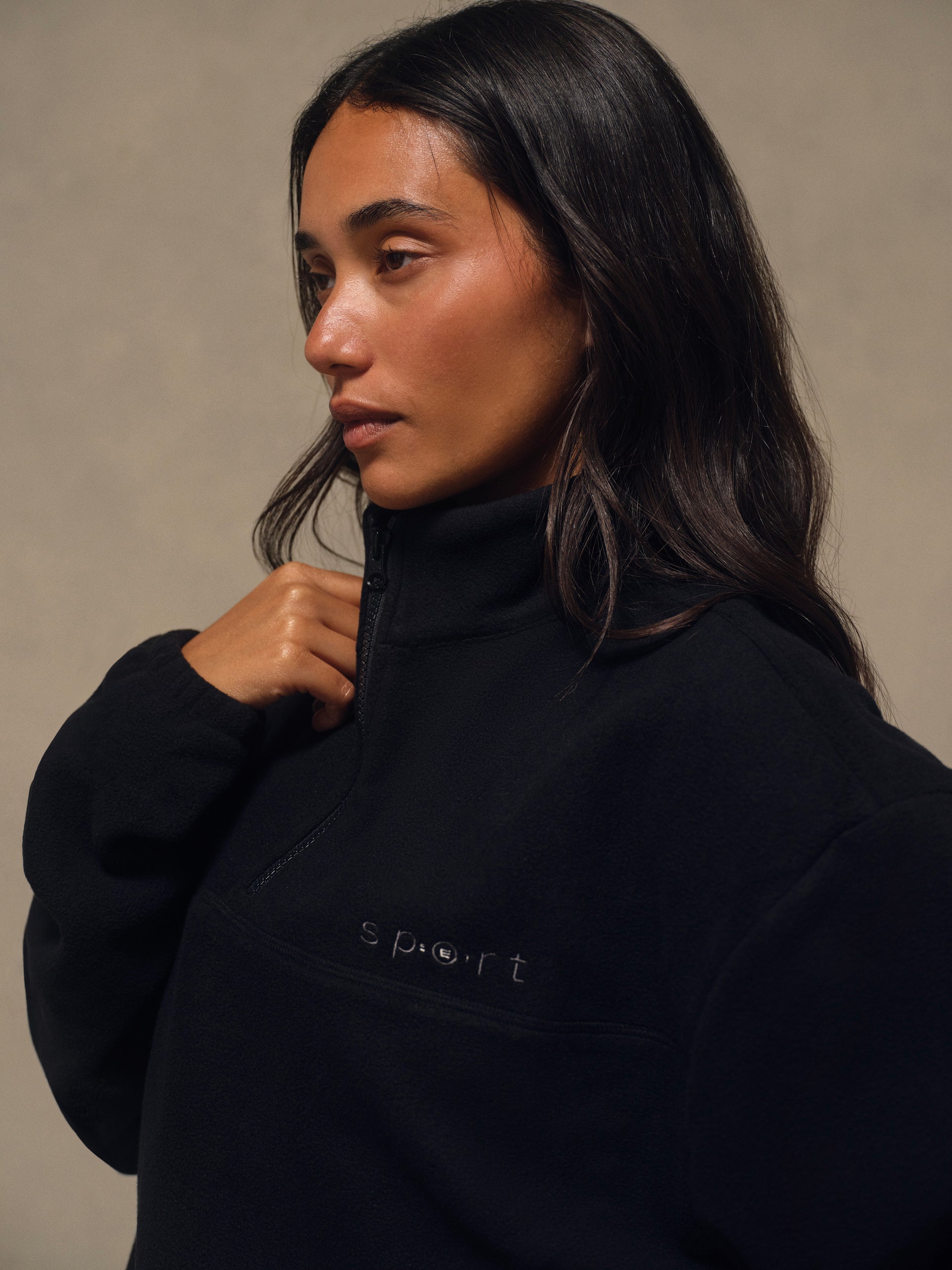 A woman with long dark hair wearing a black fleece half-zip pullover sweatshirt with 's port' embroidered on the chest, posed against a neutral beige background.
