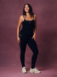A woman modeling a black sleeveless athletic jumpsuit with silver rhinestone trim along the straps and sides, paired with white sneakers, against a textured mauve-pink studio backdrop. 