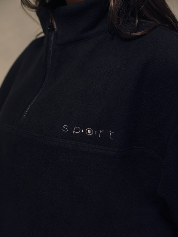 Close-up of a person wearing a dark navy quarter-zip fleece pullover with the word 'sport' embroidered on the chest.