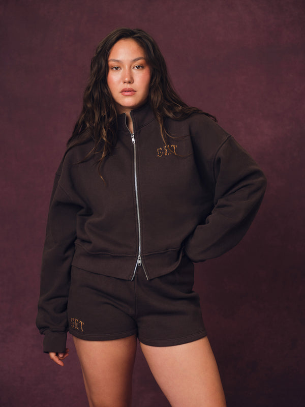 A woman modeling a dark brown zip-up sweatshirt and matching shorts set with 'SET' lettering, posed against a textured dark purple studio backdrop. 