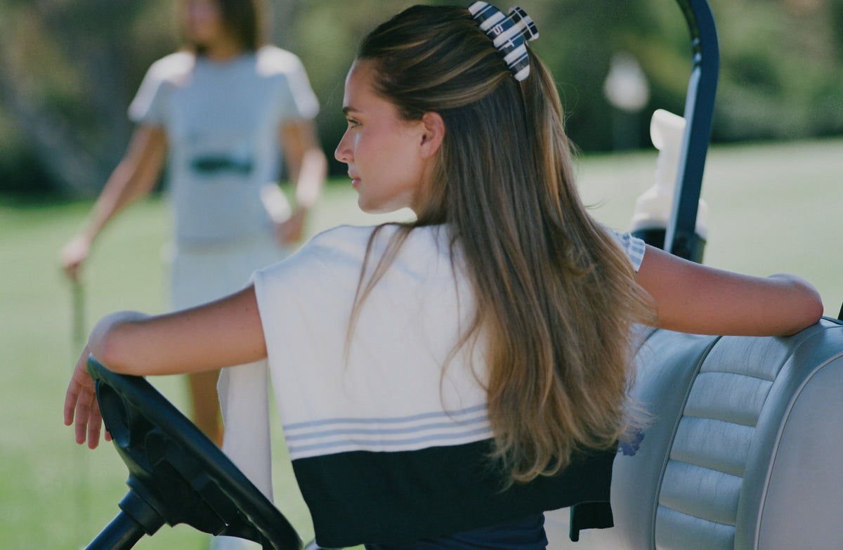A woman with long brown hair sits in the back of a golf cart on a grassy golf course, wearing a white and navy short-sleeve top with a striped hair clip. Another person is blurred in the background.
