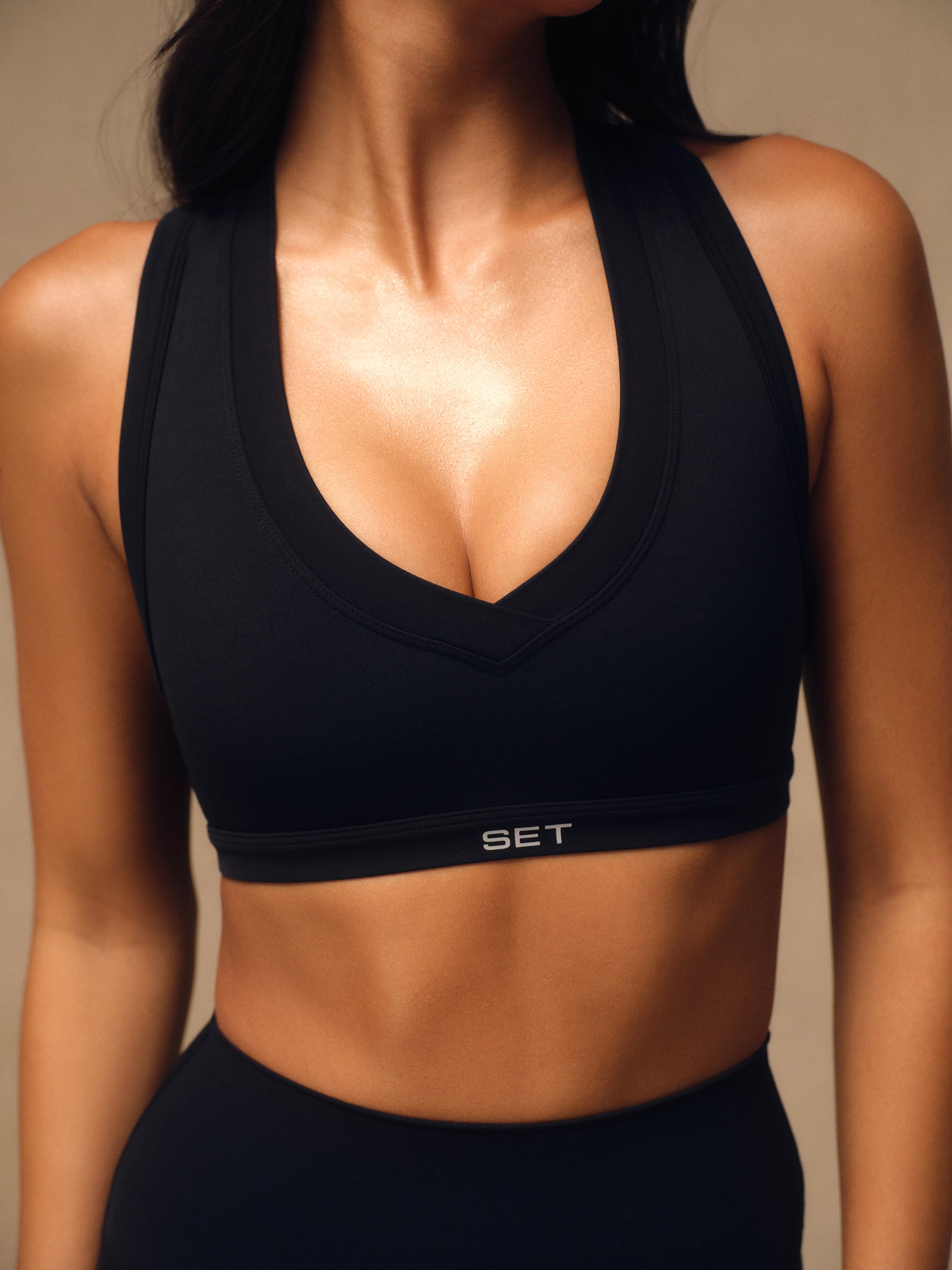 A close-up of a woman wearing a black athletic sports bra with a wide scoop neckline and the brand name 'SET' printed in white on the underband, paired with black athletic bottoms against a warm beige background. <model-info>Jelene wears a size XS and is 5’7”, 23” waist, 32AB bust.</model-info>